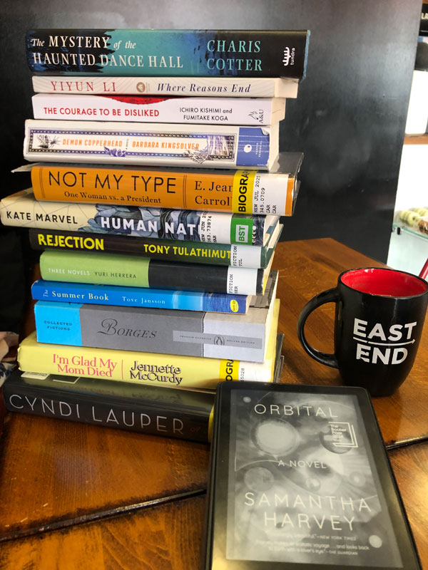 Group book pile at East Toronto Coffee Co, accompanied by coffee - titles include Not My Type by E. Jean Carroll, I’m Glad My Mom Died by Jennette McCurdy, Rejection by Tony Tulathimutte, The Courage to Be Disliked by Ichiro Kishimi and Fumitake Koga + more - Photo by Jennifer D. Foster