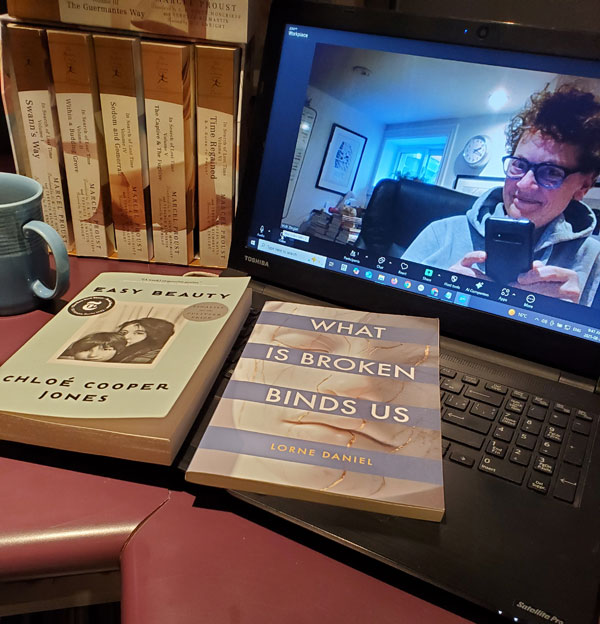 Silent book club member Vicki (that's me, with my signature messy hair and glasses), on screen getting ready for our zoom meeting, with books stacked next to my computer, including works by Marcel Proust, Chloe Cooper Jones and Lorne Daniel