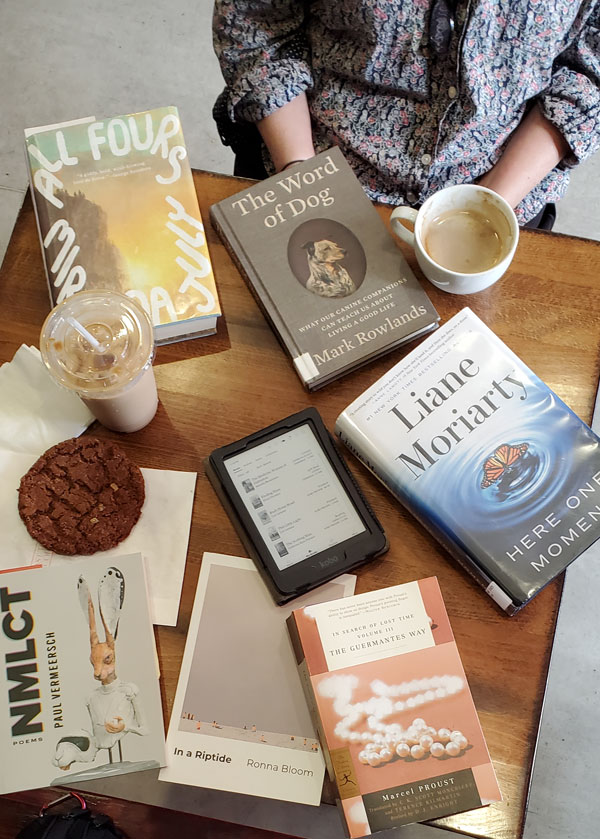 Group book pile at East Toronto Coffee Co, accompanied by coffee and pastries - titles include Here One Moment by Liane Moriarty, Volume III of In Search of Lost Time by Marcel Proust + more