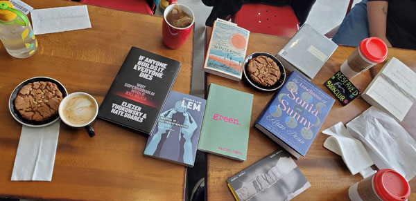 Books spread out on tables at East Toronto Coffee Co, including titles by Zachari Logan, Robin Wall Kimmerer, Adania Shibli + more, accompanied by coffee and pastries (Photo by Vicki Ziegler)