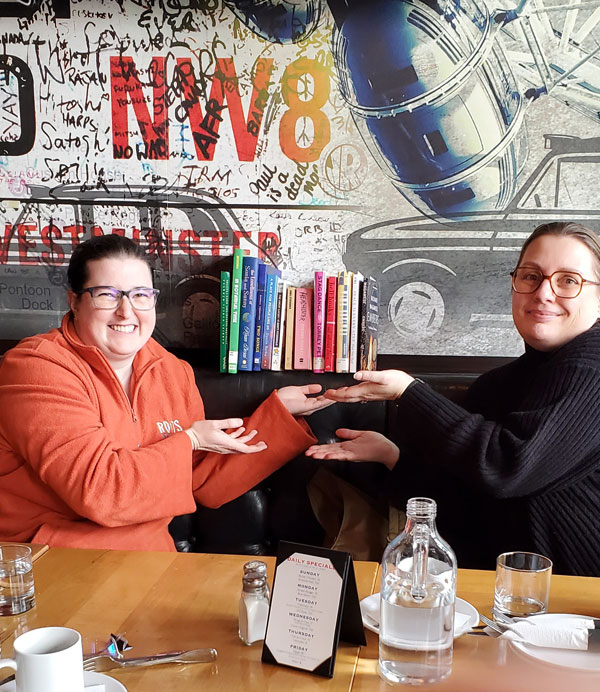 Silent book club members Ruth (left) and Jenn (right) present the collected books being read by east end Toronto sbc members at the meeting at Firkin on Danforth (Photo by Vicki Ziegler)