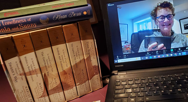 Silent book club member Vicki (oui, c'est moi) on screen, preparing for the sbc zoom meeting, with recent reading next to the computer, including the 6 volumes of In Search of Lost Time by Marcel Proust, translated by Andreas Mayor and Terence Kilmartin, revised by D.J. Enright, Green by Zachari Logan and The Loneliness of Sonia and Sunny by Kiran Desai (Photo by Vicki Ziegler)