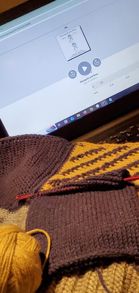 Audiobook of Marigold and Rose by Louise Glück (Farrar, Straus and Giroux) on laptop screen, with blue and muted colours crochet work sitting on laptop keyboard