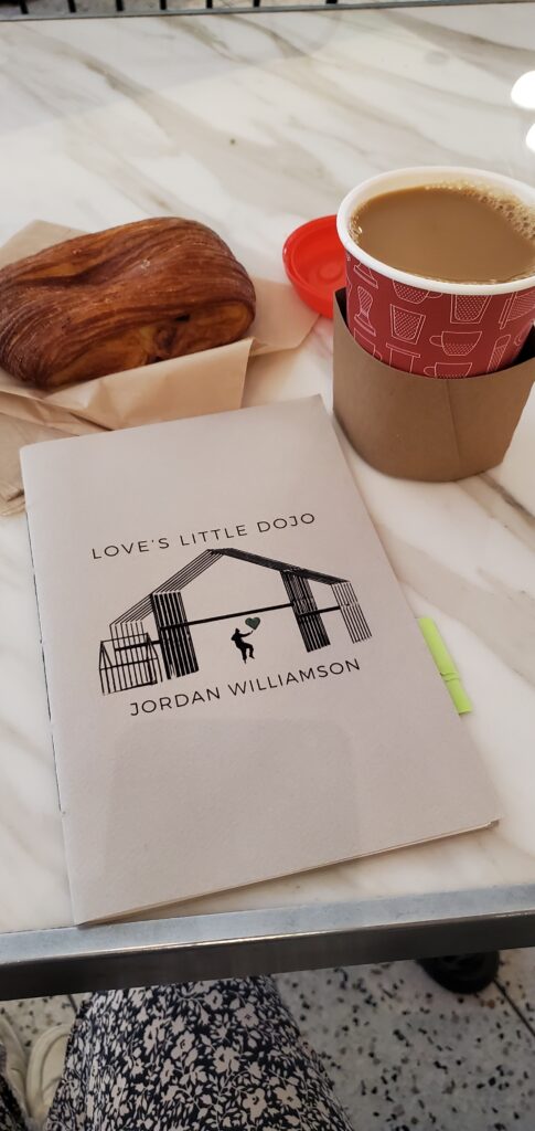 Poetry chapbook Love's Little Dojo by Jordan Williamson (Pinhole Poetry) sits on a table with a croissant and a takeout coffee