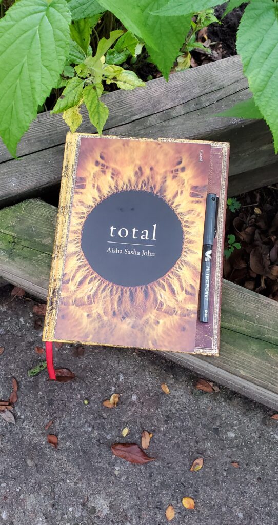 Poetry collection total by Aisha Sasha John (McClelland & Stewart) sits on old wooden garden fence with green leaves above it, along with a notebook with an ornate gold cover and a black pen