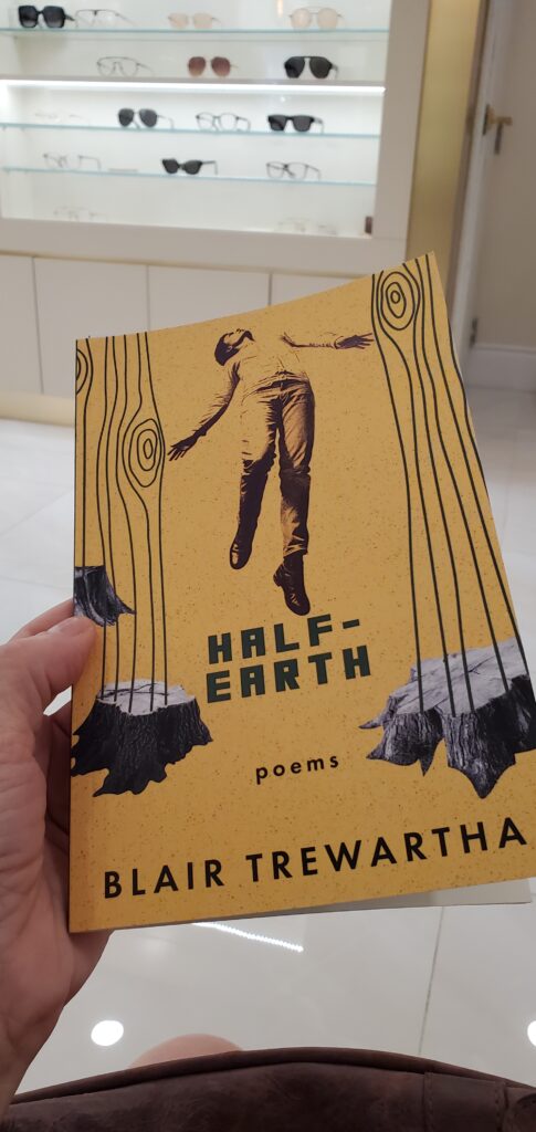 Poetry collection Half-Earth by Blair Trewartha (Palimpsest Press/Anstruther Books) being read in an optometrist's office, with shelves with eyeglasses on them in the background