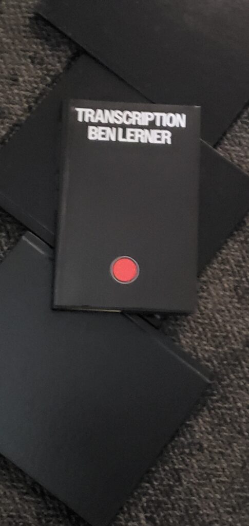Blurry shot of Transcription by Ben Lerner, with its stark black cover with a bright red dot on it, sits atop some black-covered notebooks on a gray carpet