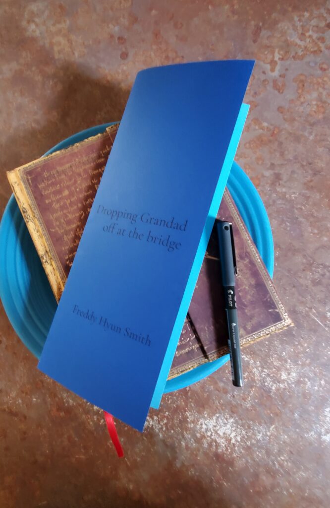 Bright blue poetry pamphlet Dropping Grandad off at the bridge by Freddy Hyun Smith (Opaat Press) sits on a blue plate with a notebook with an ornate gold cover and a black fine tip pen