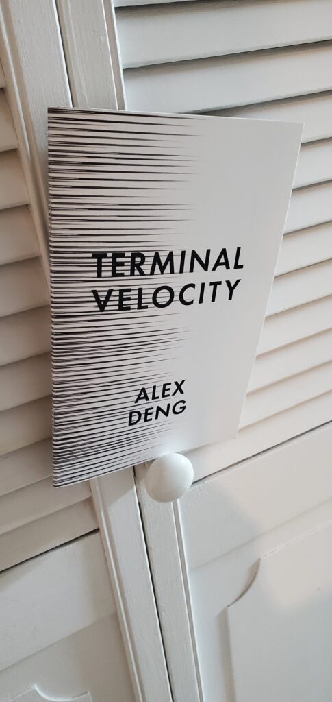Poetry chapbook Terminal Velocity by Alex Deng (Anstruther Press), with its striking black and white cover, perches on the doorknob of a door with white slats
