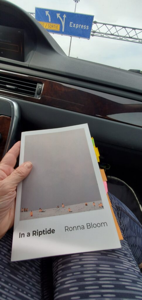 I'm holding the poetry collection In a Riptide by Ronna Bloom (Brick Books) in my lap sitting in the passenger seat of a car. Colourful post-it notes jut out of the book. Above the windshield is a traffic sign with the word 