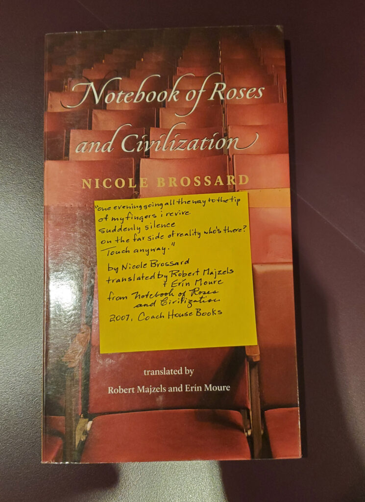 Poetry collection Notebook of Roses and Civilization by Nicole Brossard, translated by Robert Majzels and Erin Moure (Coach House Books), with its vivid red cover, with a yellow post-it note affixed in the middle with a handwritten transcription of a fragment of one of her/their poems/translations