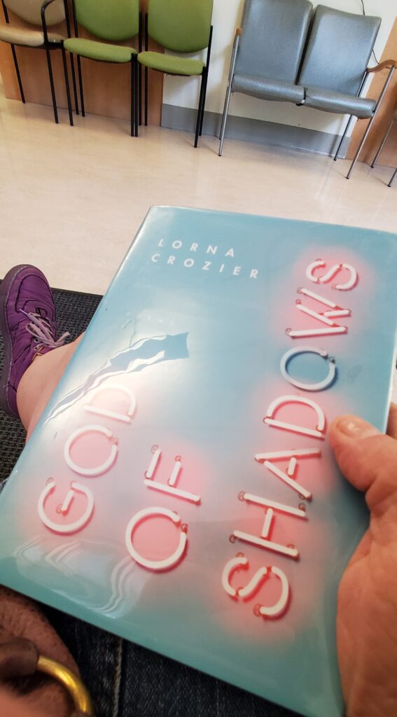 My pink/purple shoes are visible as I read God of Shadows by Lorna Crozier (McClelland & Stewart) in a waiting room