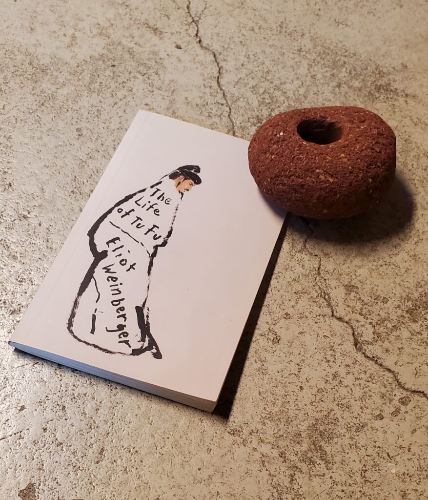 Poetry collection The Life of Tu Fu by Eliot Weinberger (New Directions) sits on a concrete floor with a round piece of red brick sitting nearby