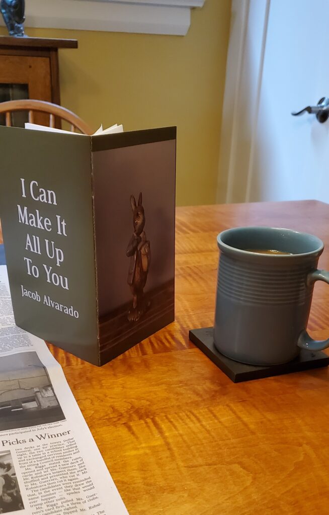 Poetry chapbook I Can Make It All Up To You by Jacob Alvarado (knife fork book) stands upright next to a blue cup of coffee