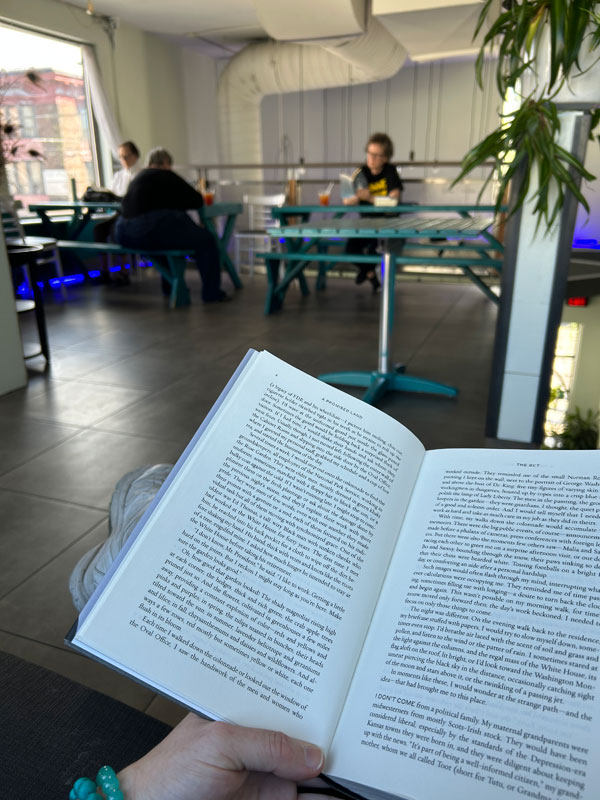 Silent book club members reading on the 2nd floor of the Sprouty Cafe, with member Ruth holding her book in the foreground (Photo by Ruth Bartlett)